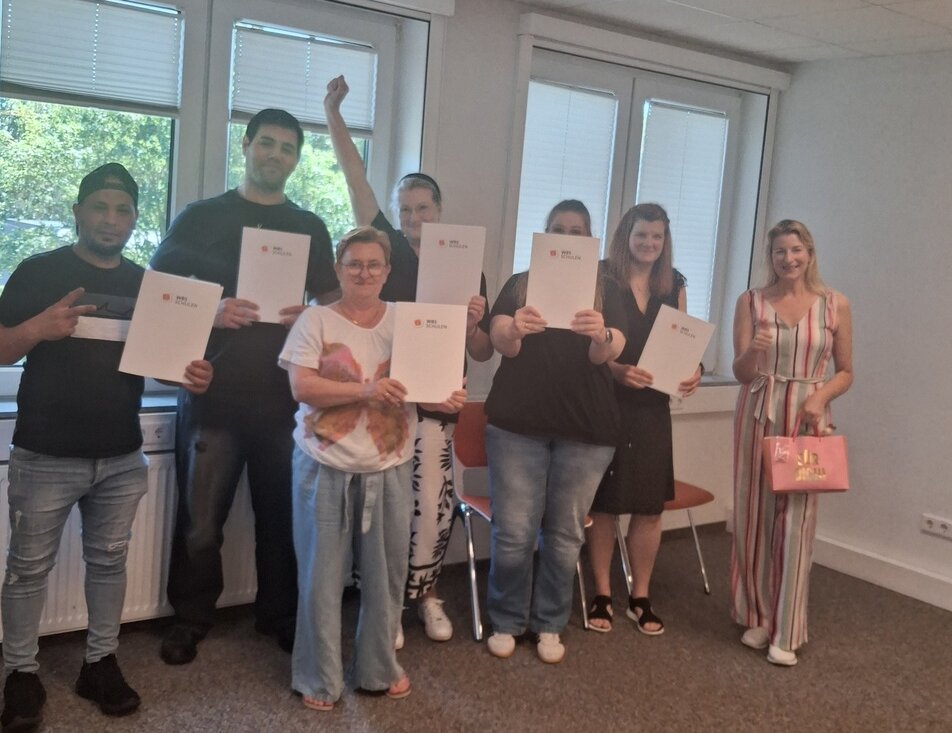 Abschlussfoto einer Schulungsgruppe mit Zertifikatsmappen in einem hellen Seminarraum.