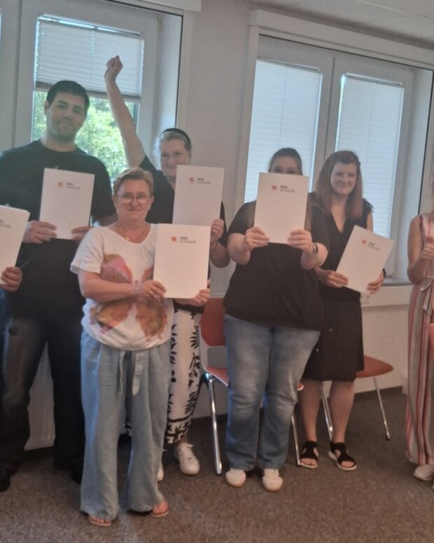 Abschlussfoto einer Schulungsgruppe mit Zertifikatsmappen in einem hellen Seminarraum.
