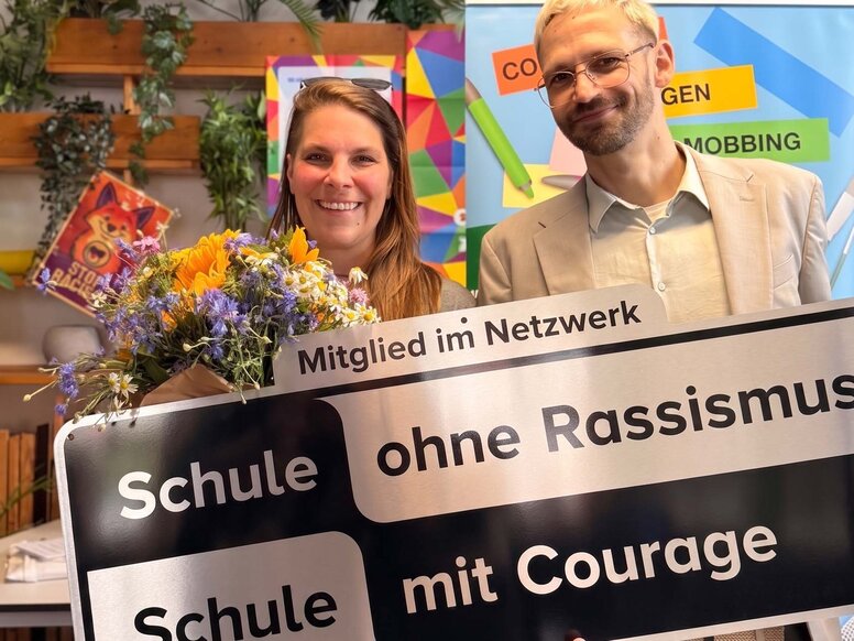 Zwei Personen aus dem Team der WBS SCHULEN Berlin präsentieren in Berlin das Schild „Schule ohne Rassismus – Schule mit Courage“. Die Auszeichnung unterstreicht das Engagement für Toleranz, Vielfalt und ein respektvolles Miteinander.