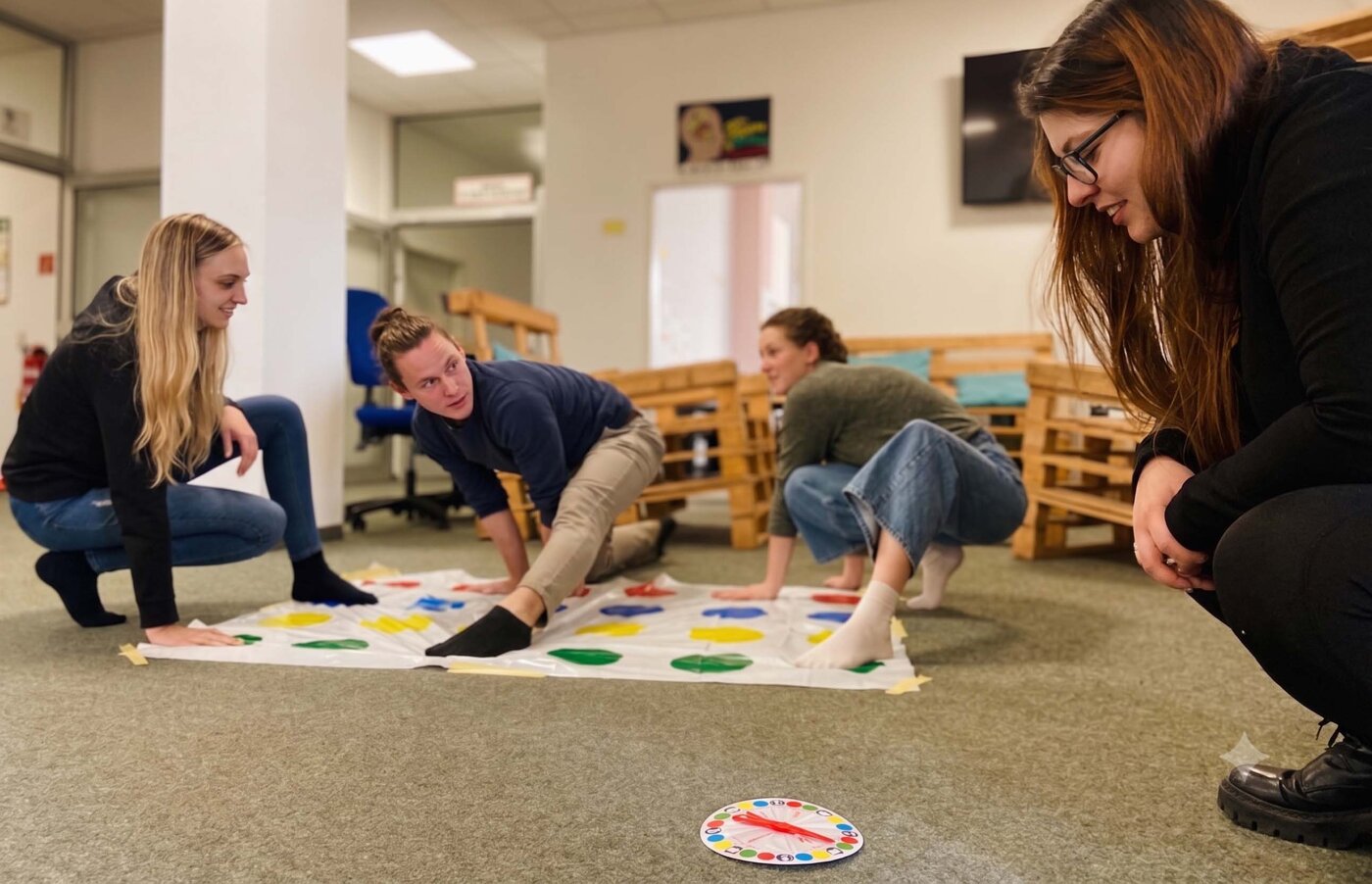 Vier junge Auszubildende spielen lachend Twister auf dem Boden in einem Aufenthaltsraum während der "gesunden Woche". Eine Person kniet und schaut zu den anderen, die sich über die bunte Matte strecken.