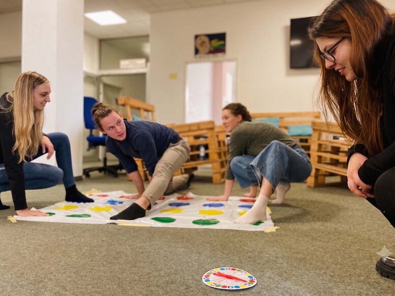 Vier junge Auszubildende spielen lachend Twister auf dem Boden in einem Aufenthaltsraum während der "gesunden Woche". Eine Person kniet und schaut zu den anderen, die sich über die bunte Matte strecken.