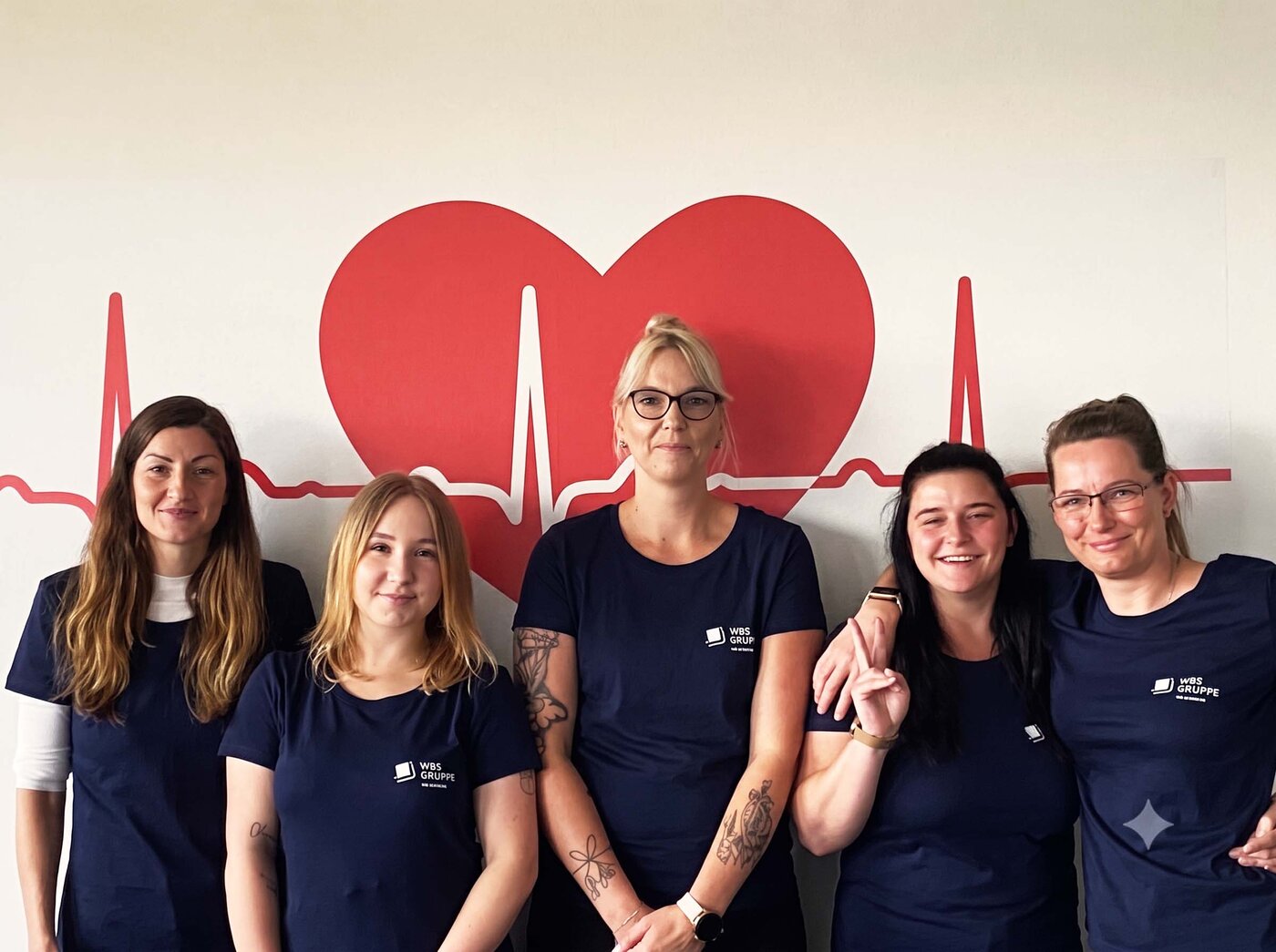Fünf Mitglieder des Teams WBS SCHULEN Magdeburg posieren in Laufshirts vor einer Wand mit Herz- und EKG-Motiv. Das Foto entstand bei der Vorbereitung auf den Firmenstaffellauf.