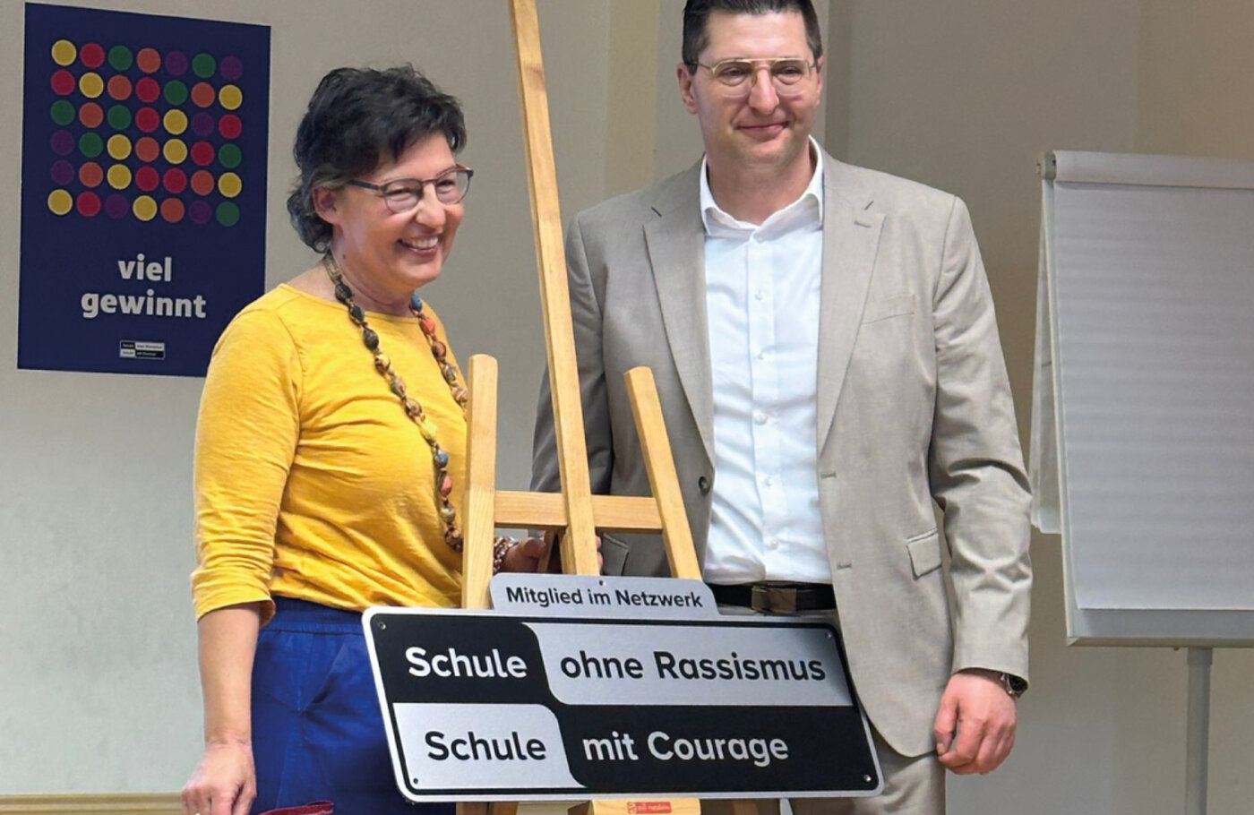 Schulleitung und Bürgermeister stehen vor einer Staffelei mit dem Siegel „Schule ohne Rassismus – Schule mit Courage“ bei der feierlichen Übergabe in einer Schule in Plauen und lächeln.