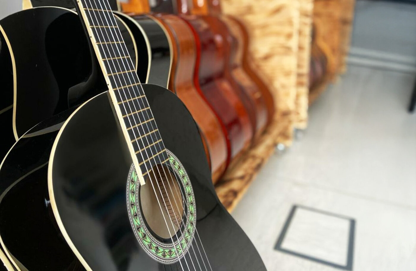 Nahaufnahme einer schwarzen Akustikgitarre mit heller Umrandung am Schallloch. Im Hintergrund stehen mehrere weitere, holzfarbene Gitarren aufgereiht in einem Holzständer.
