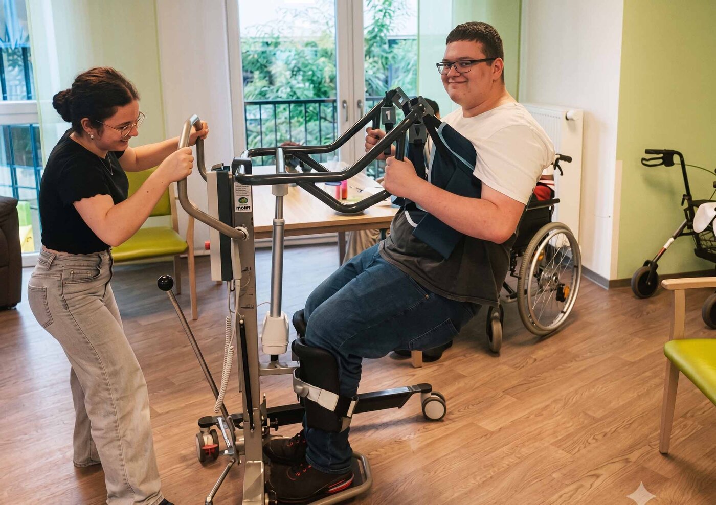 Sozialassistent:innen üben im Pflegeeinsatz die Transfertechnik mit einem Patientenlifter. Eine Person wird sicher vom Rollstuhl umgesetzt, während eine andere Person die Durchführung begleitet.