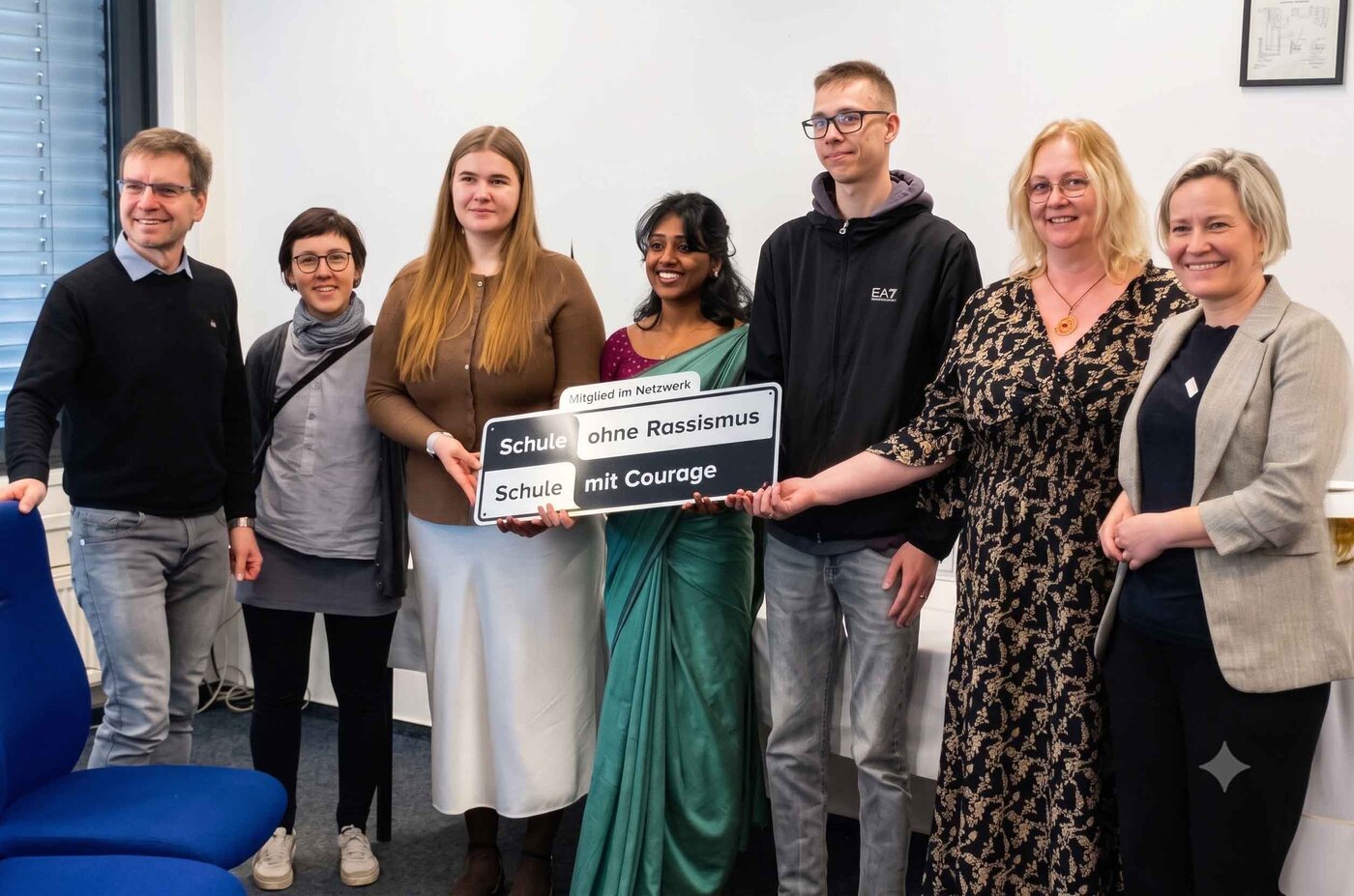 Ein Gruppenfoto von sieben Personen in einem hellen Büroraum, die gemeinsam ein Schild hochhalten. Das Schild zeigt oben den Text "Mitglied im Netzwerk" und darunter die Logos und Texte "Schule ohne Rassismus" (links) und "Schule mit Courage" (rechts).