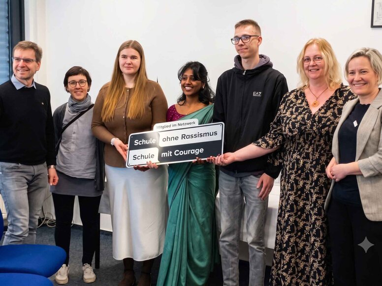 Ein Gruppenfoto von sieben Personen in einem hellen Büroraum, die gemeinsam ein Schild hochhalten. Das Schild zeigt oben den Text "Mitglied im Netzwerk" und darunter die Logos und Texte "Schule ohne Rassismus" (links) und "Schule mit Courage" (rechts).