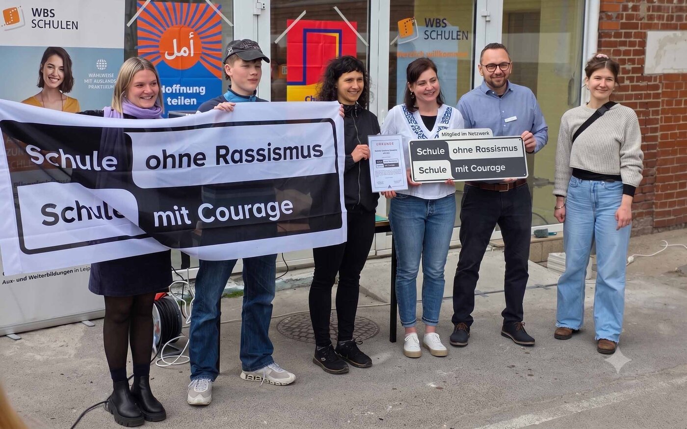 Sechs Personen halten vor dem Schulgebäude der WBS SCHULEN Leipzig ein Banner und Schild mit "Schule ohne Rassismus - Schule mit Courage". Personen zeigen Zertifikate.