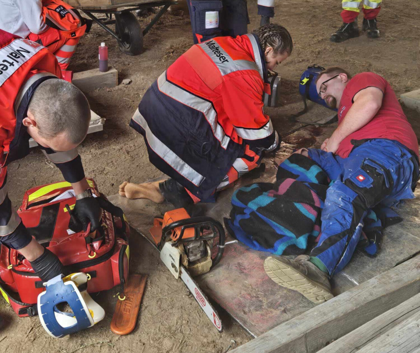 Mehrere Personen in Rettungsdienstkleidung versorgen bei einer Übung in einer Scheune einen Mann mit einer schweren Beinverletzung. Medizinische Ausrüstung liegt am Boden, daneben eine Kettensäge.