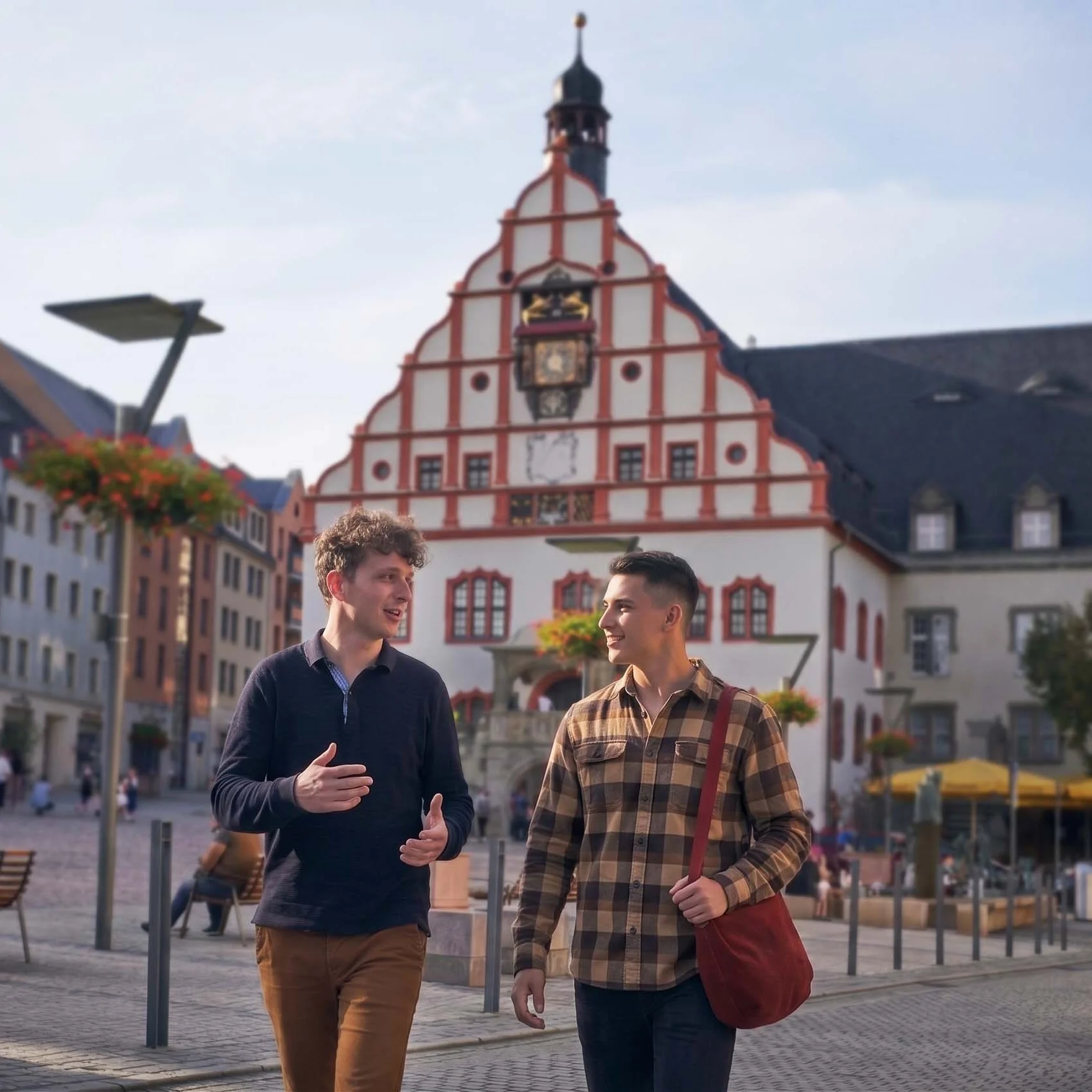 Zwei junge Männer unterhalten sich, während sie über den Altmarkt in Plauen gehen. Im Hintergrund ist das alte Rathaus mit Rathausturm zu sehen.