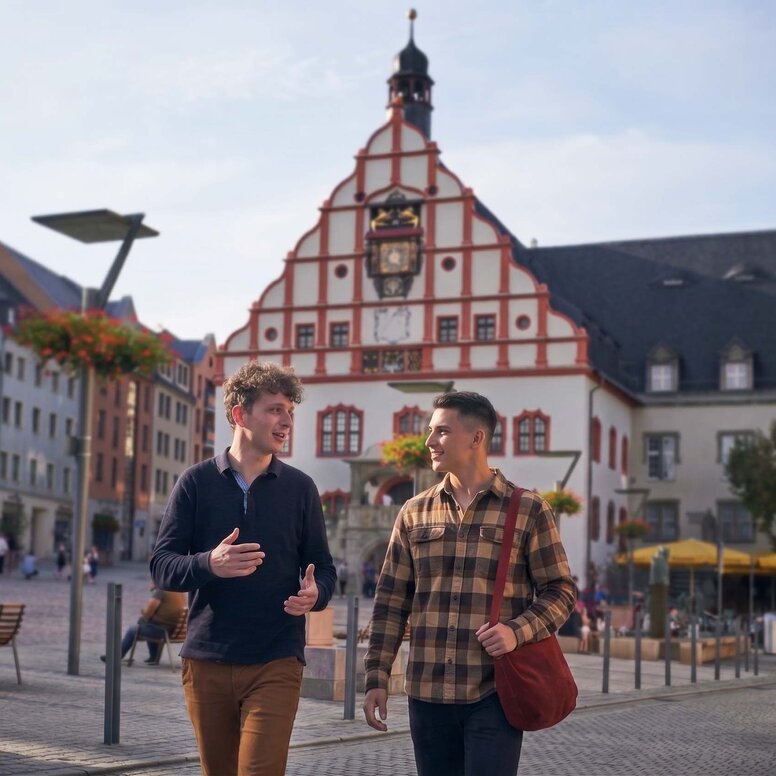 Zwei junge Männer unterhalten sich, während sie über den Altmarkt in Plauen gehen. Im Hintergrund ist das alte Rathaus mit Rathausturm zu sehen.