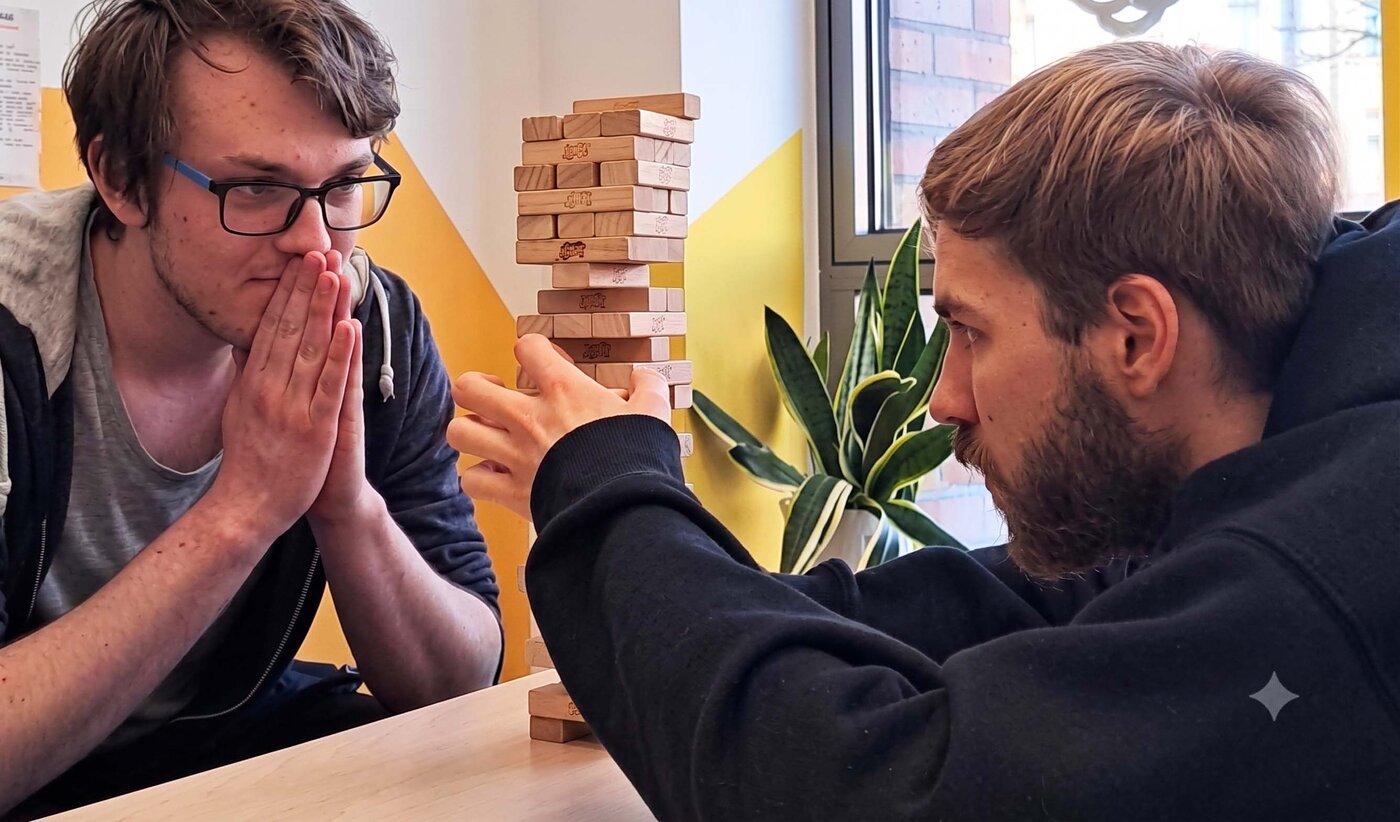 Zwei Auszubildende spielen während des praktischen Unterrichts konzentriert Jenga an einem Holztisch. Der junge Mann rechts im Hoodie zieht vorsichtig einen Holzblock heraus.