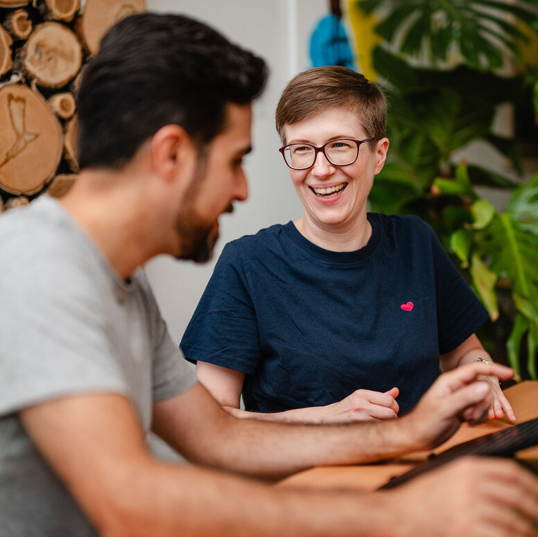 Zwei Mitarbeitenden sitzen an einem Tisch, lachen und arbeiten am Laptop. 