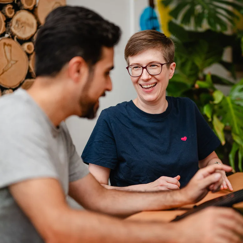 Zwei Mitarbeitenden sitzen an einem Tisch, lachen und arbeiten am Laptop. 