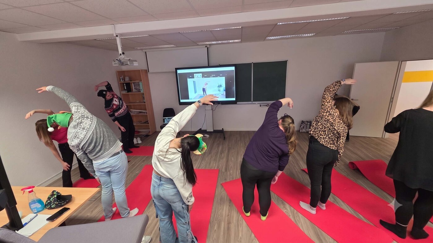 Mehrere Personen führen auf roten Yogamatten Dehnübungen in einem Schulungsraum durch. Vorne zeigt ein Bildschirm eine angeleitete Übung, während die Teilnehmenden die seitliche Streckbewegung nachmachen.