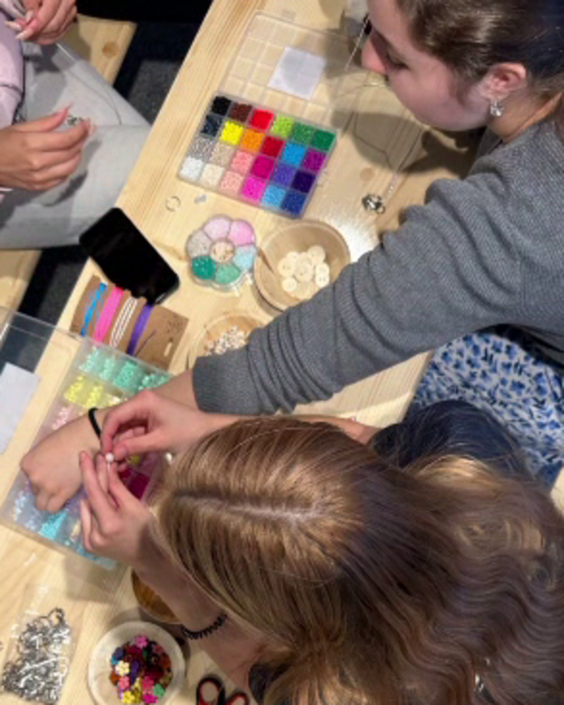 Auf einem Holztisch gestalten mehrere Personen gemeinsam Schmuck oder Bastelarbeiten. Auf dem Tisch liegen Perlen, Anhänger, Scheren, Schnüre und Sortierboxen mit buntem Bastelmaterial, während mehrere Hände an den Materialien arbeiten.
