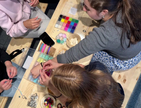 Auf einem Holztisch gestalten mehrere Personen gemeinsam Schmuck oder Bastelarbeiten. Auf dem Tisch liegen Perlen, Anhänger, Scheren, Schnüre und Sortierboxen mit buntem Bastelmaterial, während mehrere Hände an den Materialien arbeiten.