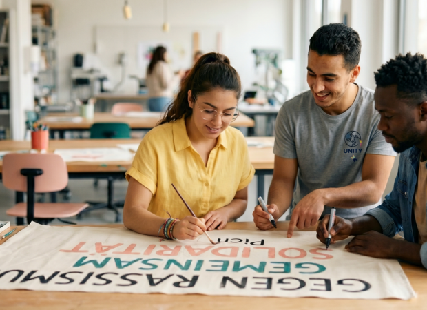 Diverse Studenten malen engagiert an einem Plakat gegen Rassismus in einem hellen Atelier, als Teil eines praxisorientierten sozialen Projektes zur Solidarität.