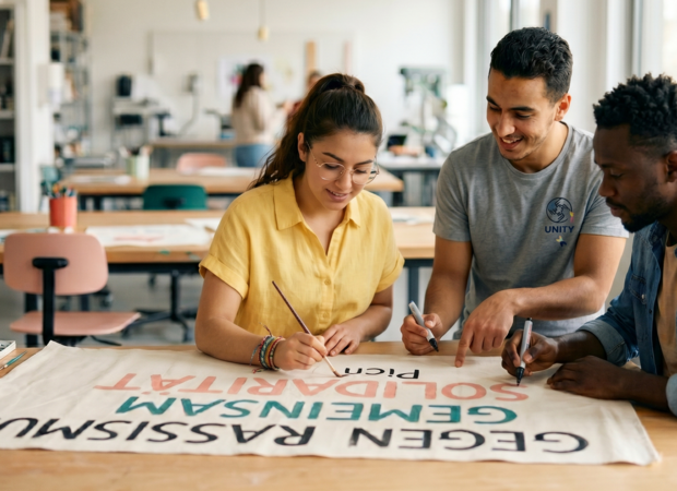 Diverse Studenten malen engagiert an einem Plakat gegen Rassismus in einem hellen Atelier, als Teil eines praxisorientierten sozialen Projektes zur Solidarität.