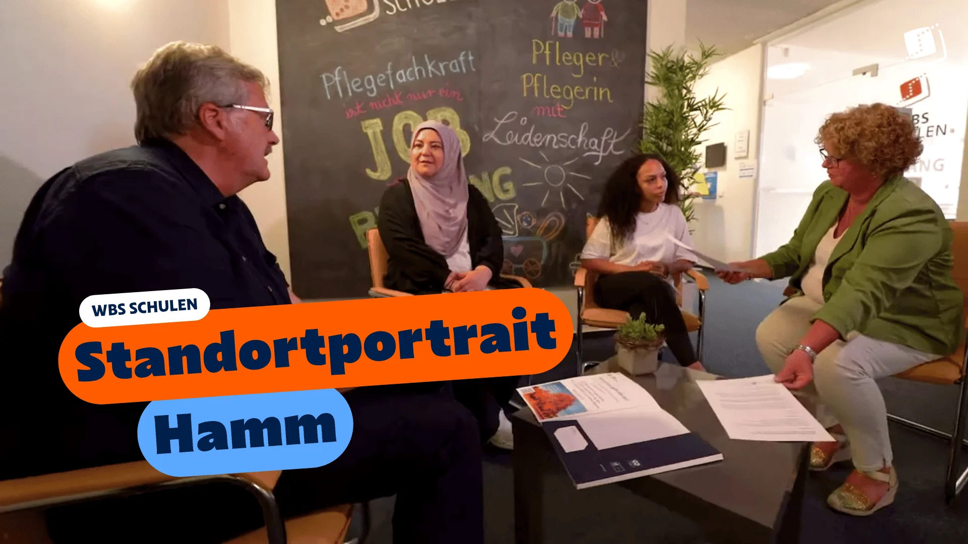 Vier Personen sitzen in einem Besprechungsbereich und sprechen miteinander. Im Vordergrund liegen Unterlagen auf einem Tisch, im Hintergrund ist eine beschriftete Tafel zu sehen. Eingeblendeter Text: „WBS SCHULEN – Standortportrait Hamm“.