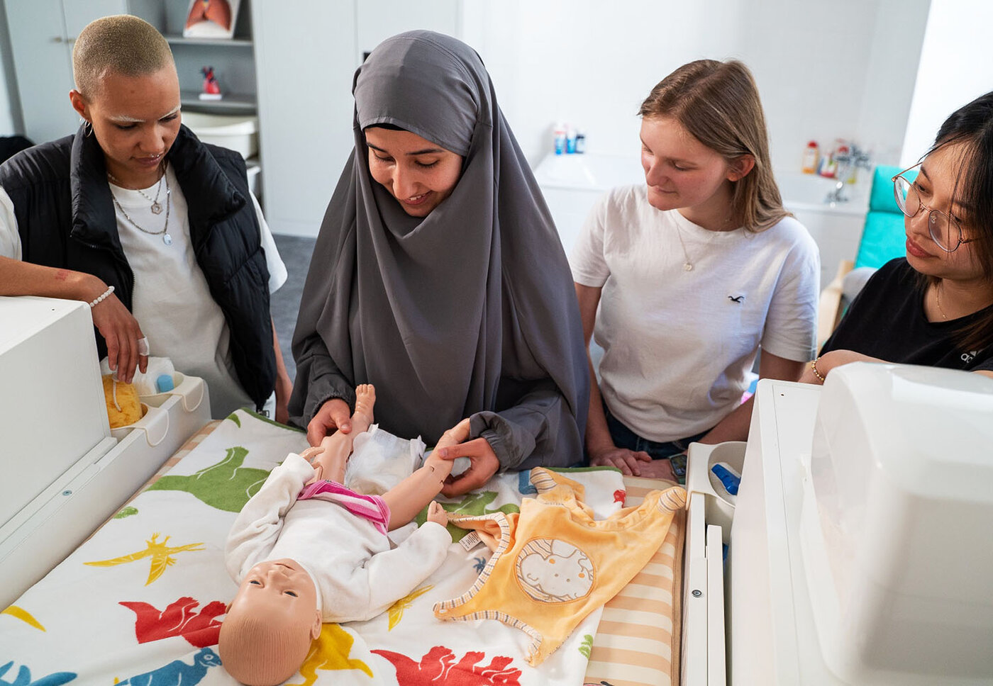 Vier Lernende stehen gemeinsam um einen Wickeltisch herum. Eine Lernende in der Mitte zieht einer lebensechten Babypuppe eine Windel aus. Die anderen drei schauen dabei aufmerksam zu.