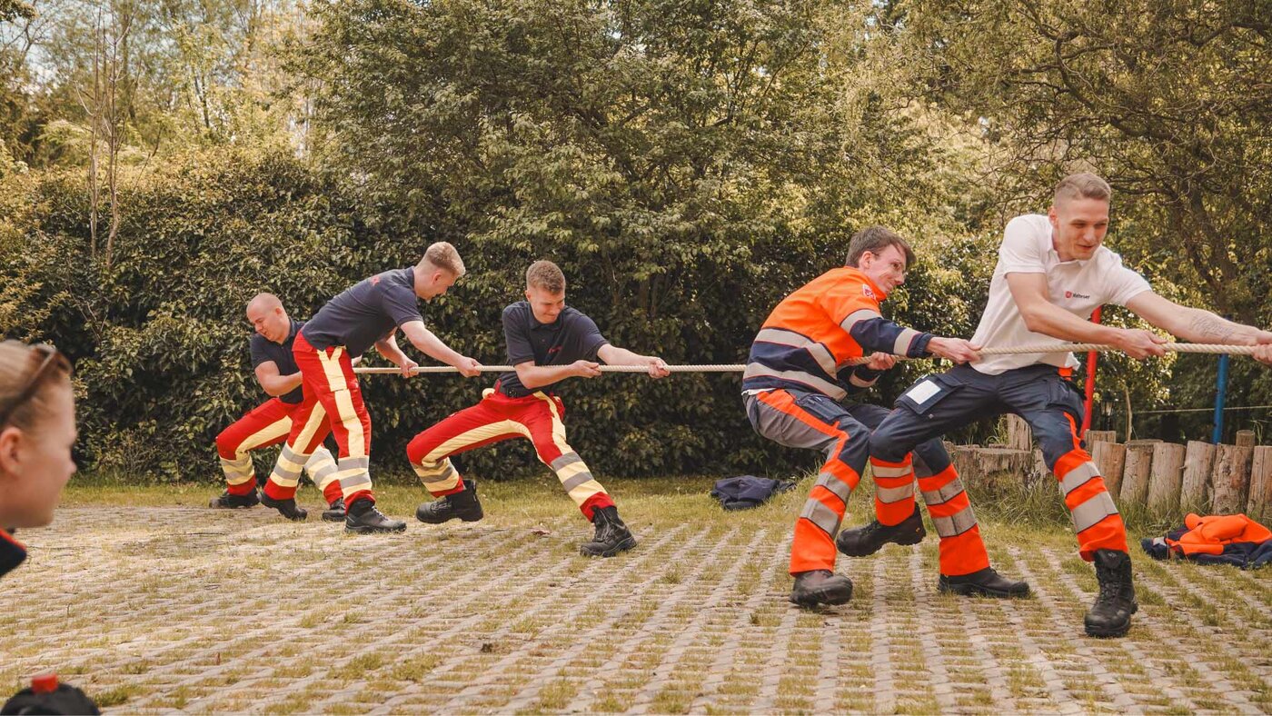 Mehrere Personen in Rettungsdienstkleidung nehmen im Freien an einem Tauzieh-Wettbewerb teil. Die Teilnehmenden ziehen gemeinsam an einem Seil und demonstrieren Teamarbeit und körperlichen Einsatz.