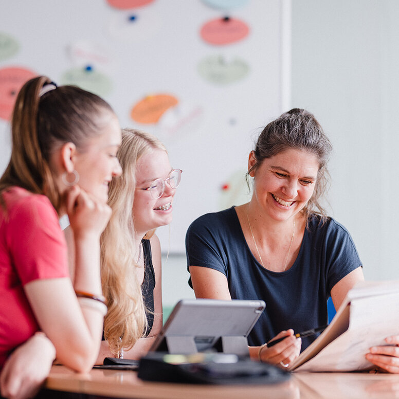 Eine Lernbegleiterin sitzt mit zwei Schülerinnen an einem Tisch in einem Klassenraum der WBS SCHULEN Berlin und schauen gemeinsam auf die Unterrichtsmaterialien.