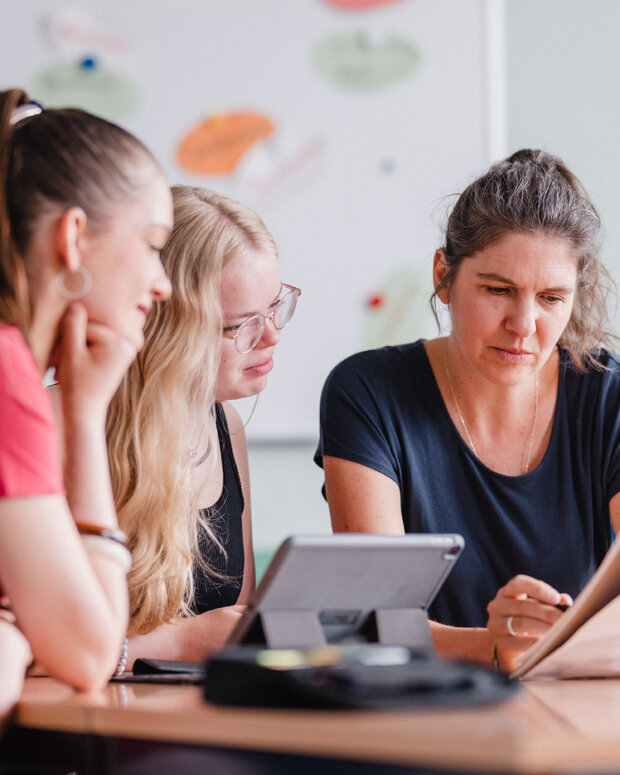 Zwei Schülerinnen und eine Lernbegleitung sitzen an einem Besprechungstisch und betrachten gemeinsam Unterlagen sowie ein Tablet. Im Hintergrund ist ein Whiteboard mit farbigen Moderationskarten zu sehen.