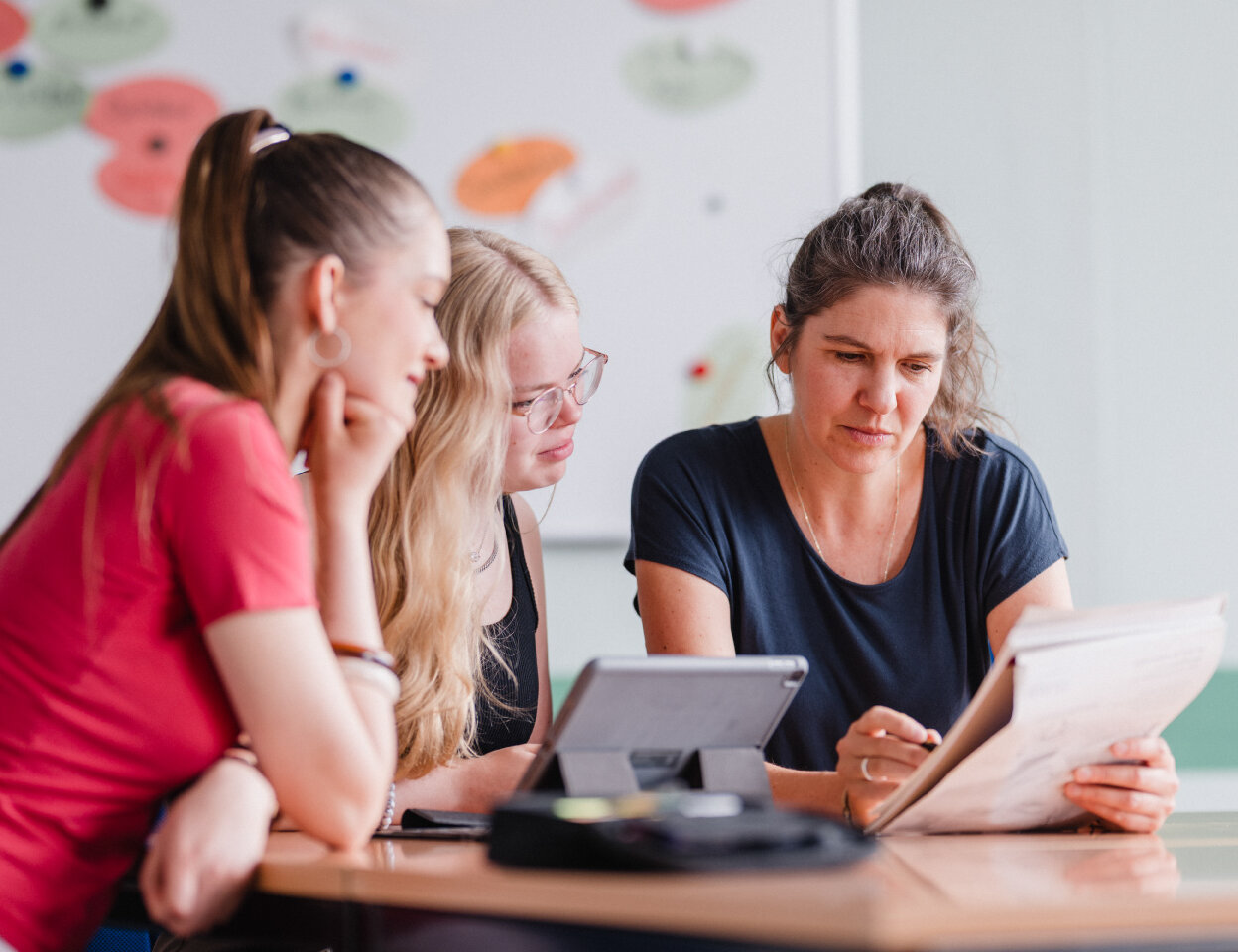 Zwei Schülerinnen und eine Lernbegleitung sitzen an einem Besprechungstisch und betrachten gemeinsam Unterlagen sowie ein Tablet. Im Hintergrund ist ein Whiteboard mit farbigen Moderationskarten zu sehen.