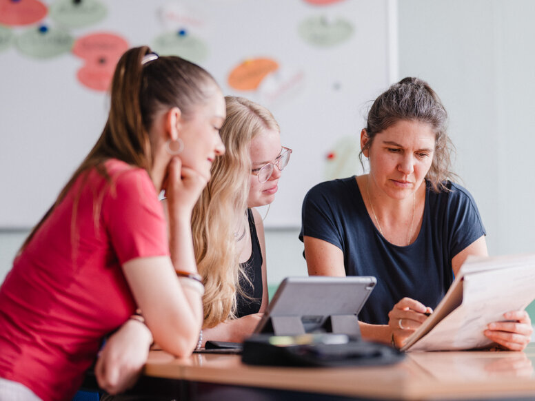 Zwei Schülerinnen und eine Lernbegleitung sitzen an einem Besprechungstisch und betrachten gemeinsam Unterlagen sowie ein Tablet. Im Hintergrund ist ein Whiteboard mit farbigen Moderationskarten zu sehen.