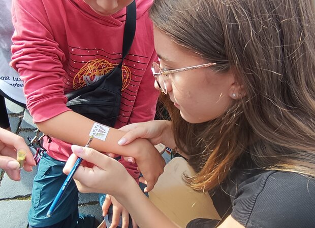 Mehrere Schülerinnen und Schüler gestalten an einem Stand im Freien temporäre Tattoos auf einem Unterarm. Im Vordergrund wird mit einem Pinsel ein Motiv aufgetragen, im Hintergrund ist eine Schreibmaschine auf dem Tisch zu sehen.