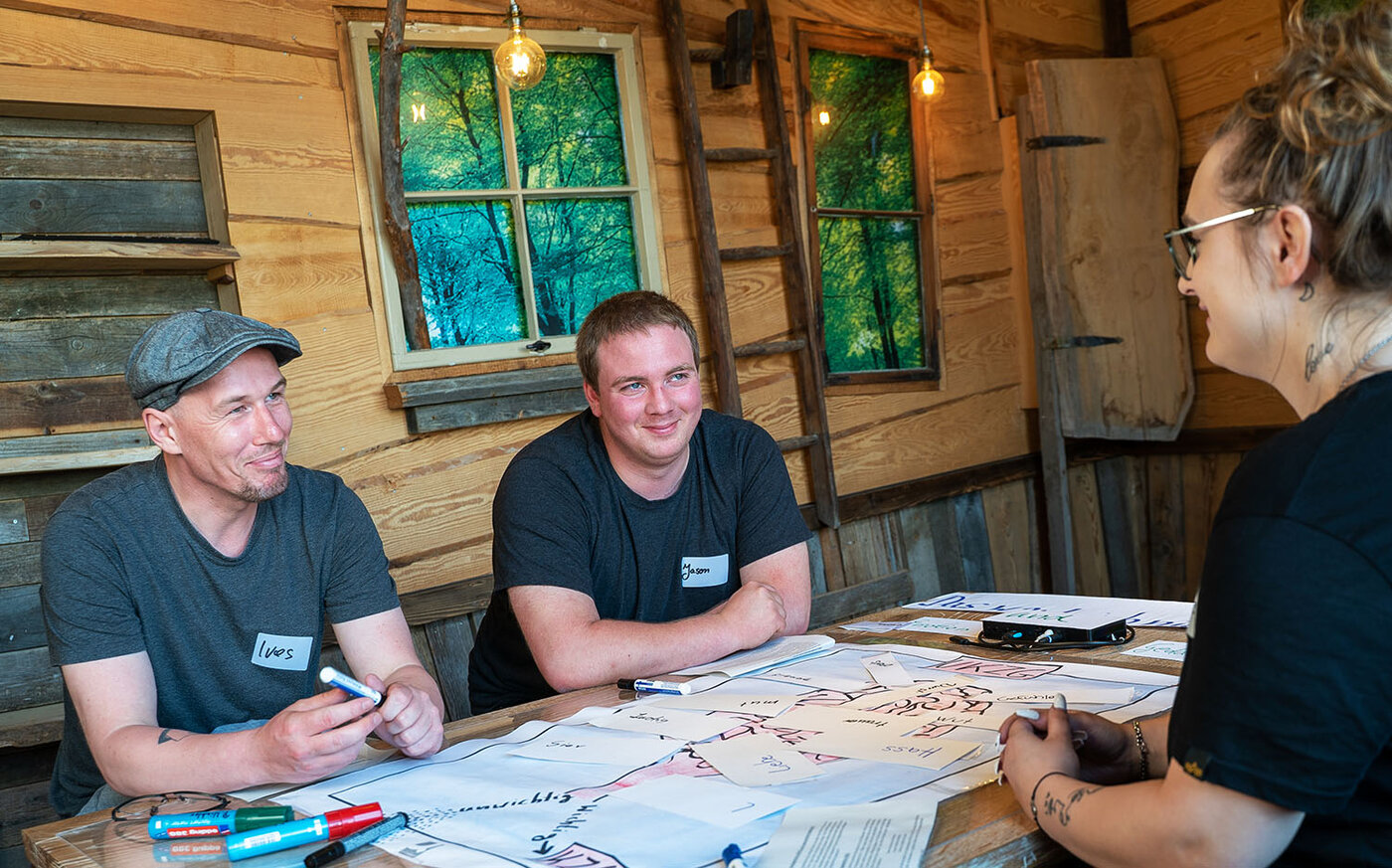 Drei Lernende sitzen an einem Tisch in einem Raum mit Holzwänden. Sie betrachten verschiedene beschriftete Papierkarten, die auf einem großen Bogen Papier liegen, und halten Textmarker in den Händen.