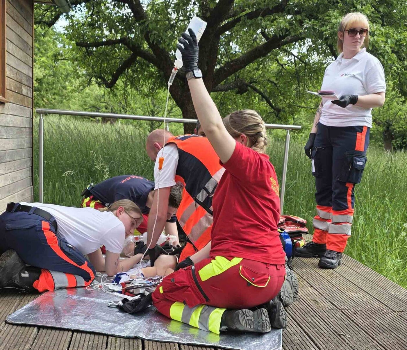Mehrere Personen in Rettungsdienstkleidung versorgen bei einer Praxisübung im Freien ein liegendes Kindermodell. Eine Person hält einen Infusionsbeutel, während andere die medizinische Versorgung auf einer Holzplattform durchführen.