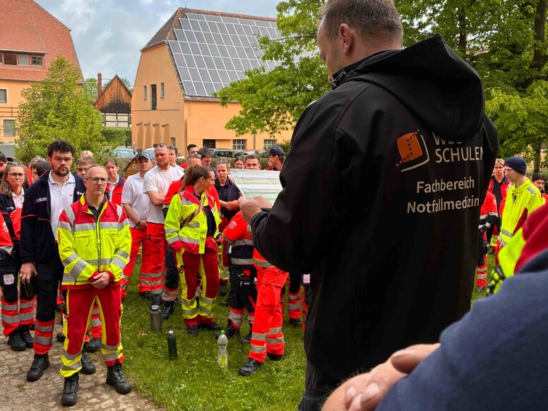 Gruppe von Auszubildenden und Einsatzkräften in rot-gelber Notfallkleidung bei einer Einsatzbesprechung. Im Vordergrund steht eine Person mit einer schwarzen Jacke mit der Aufschrift „WBS SCHULEN – Fachbereich Notfallmedizin“ und liest von einem Dokument vor. Die Szene findet im Außenbereich zwischen Bäumen und Gebäuden statt.
