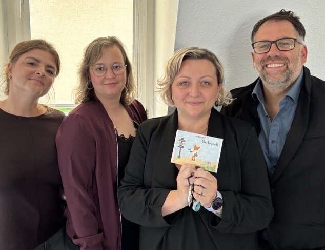 Gruppenfoto vom Schulteam in der Schule Ilmenau; die Person in der Mitte präsentiert eine kleine illustrierte Karte zur Eröffnung des neuen Standortes in Ilmenau.
