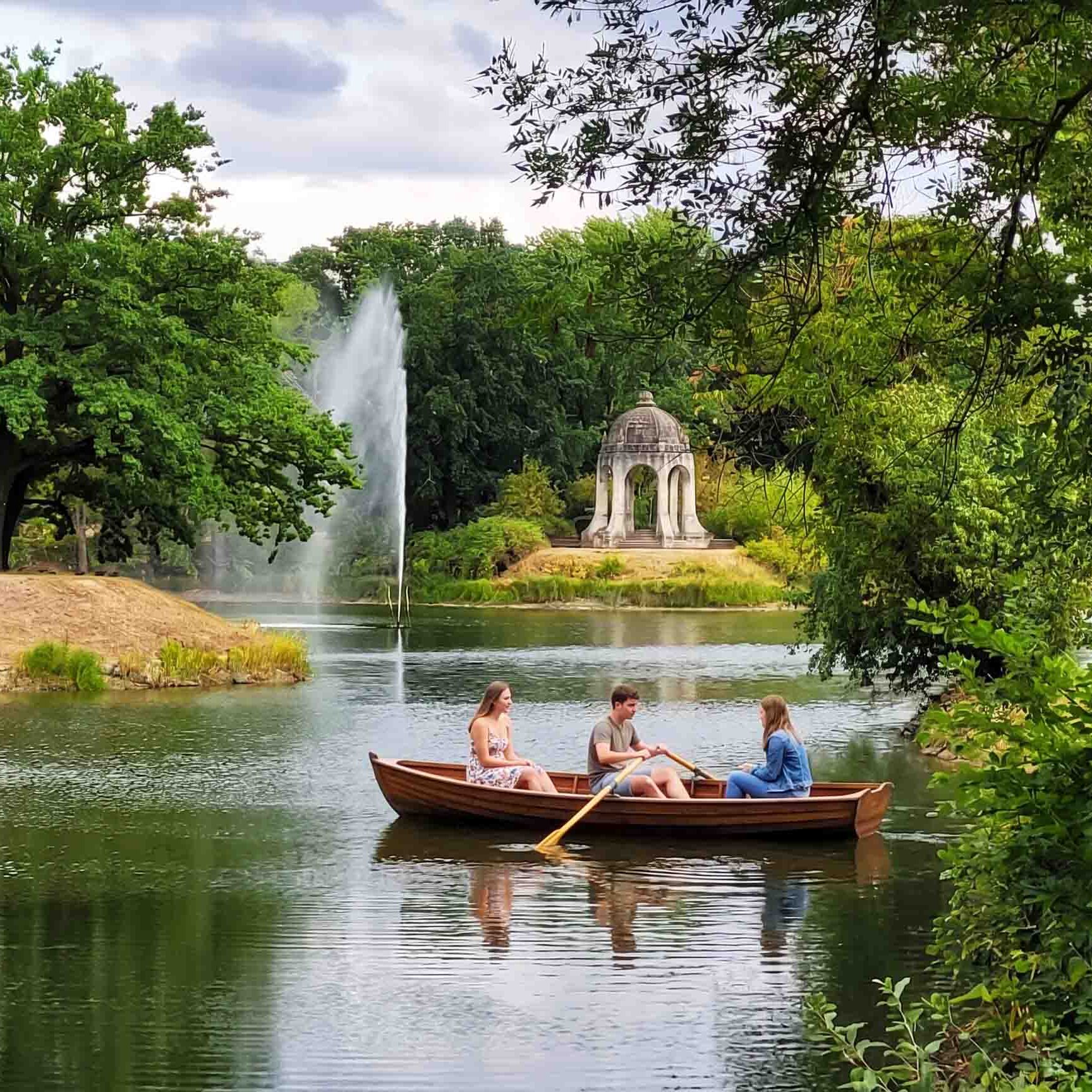 Drei Personen fahren in einem Holzruderboot über Adolf-Mittag-See in Magdeburg. Im Hintergrund sind eine hohe Wasserfontäne und der Venustempel zu sehen.
