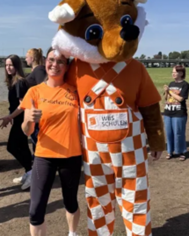 Lehrkraft in orangefarbenem T-Shirt steht auf einem Freigelände neben einer großen Fuchs-Maskottchenfigur mit dem Schriftzug „WBS SCHULEN“ auf der Hose. Mehrere weitere Personen befinden sich im Hintergrund bei einem Lauf-Event.