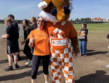Lehrkraft in orangefarbenem T-Shirt steht auf einem Freigelände neben einer großen Fuchs-Maskottchenfigur mit dem Schriftzug „WBS SCHULEN“ auf der Hose. Mehrere weitere Personen befinden sich im Hintergrund bei einem Lauf-Event.