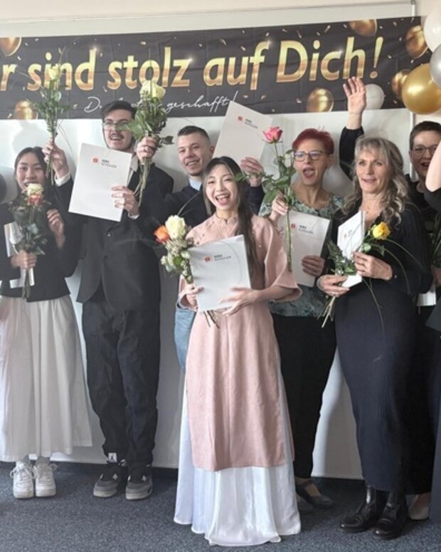 Abschlussfeier einer Pflegehilfe-Ausbildung: Absolventinnen und Absolventen präsentieren ihre Zertifikate und Blumen vor einem Gratulationsbanner.