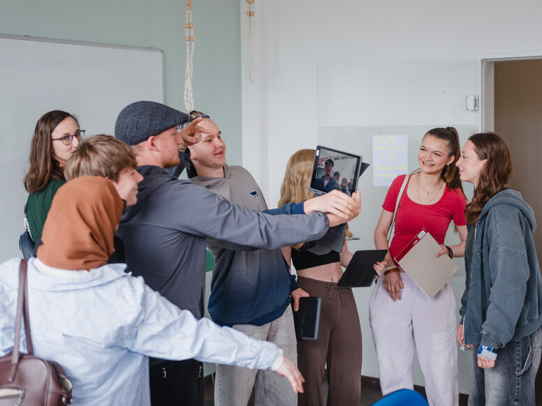 Mehrere Auszubildende stehen in einem Unterrichtsraum vor Whiteboard und Glastafel. Eine Person hält ein Tablet für ein Gruppenfoto oder einen Videoanruf, während andere Personen sich darum versammeln; einige tragen weitere Tablets oder Unterlagen.