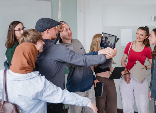 Mehrere Auszubildende stehen in einem Unterrichtsraum vor Whiteboard und Glastafel. Eine Person hält ein Tablet für ein Gruppenfoto oder einen Videoanruf, während andere Personen sich darum versammeln; einige tragen weitere Tablets oder Unterlagen.