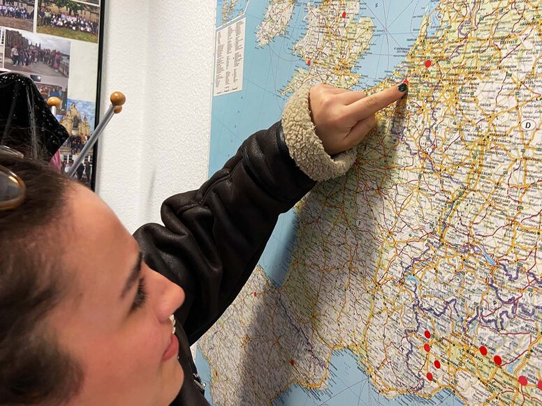 Nahaufnahme einer jungen Frau im Profil, die mit dem Finger auf eine große Europakarte an der Wand zeigt, auf der rote Markierungspunkte kleben