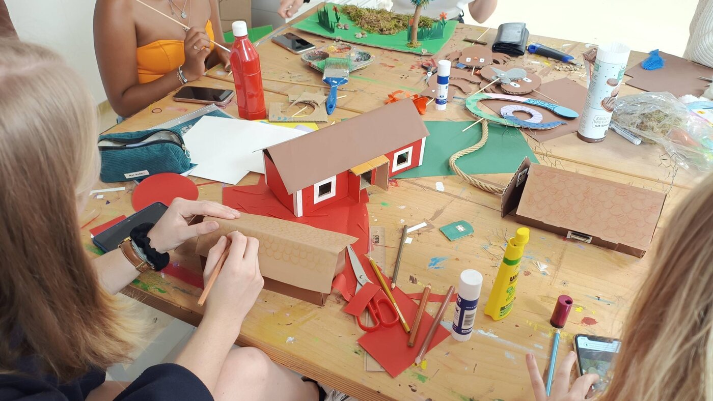 Mehrere Schüler:innen sitzen an einem Arbeitstisch und gestalten aus Karton, Papier und Naturmaterialien verschiedene Modelle, darunter kleine Häuser und Landschaften. Auf dem Tisch liegen Bastelutensilien wie Scheren, Klebstoff, Farben und Stifte.