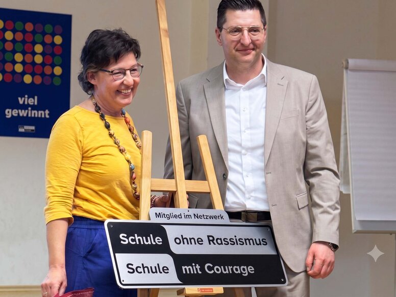 Eine Frau in gelbem Oberteil und ein Mann im hellen Anzug stehen lächelnd neben einer Holzstaffelei. Darauf steht gut sichtbar das schwarz-weiße Schild der Aktion „Schule ohne Rassismus – Schule mit Courage“