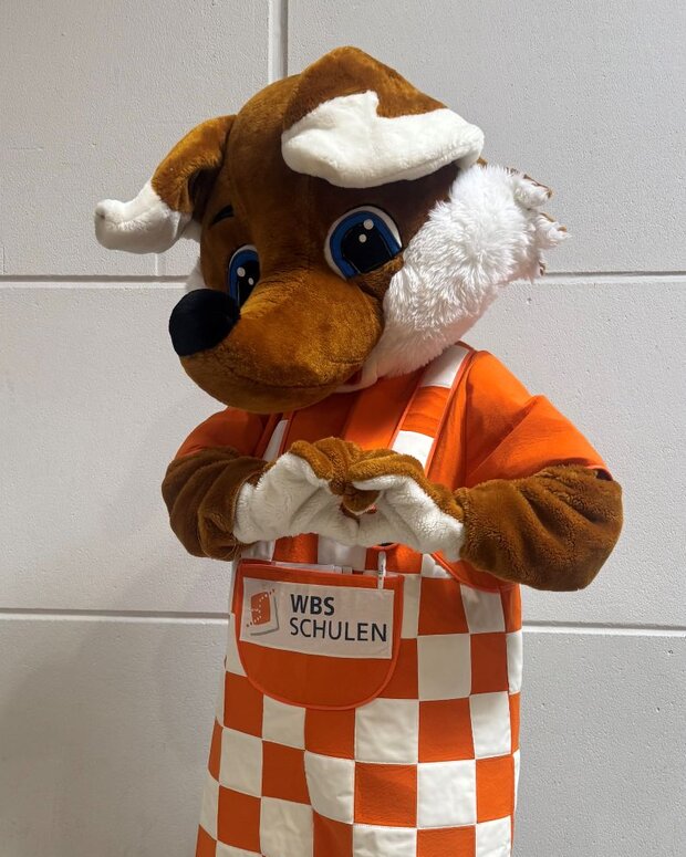 Maskottchen in Form eines Fuchses mit großem Kopf und blauem Augenpaar steht vor einer hellen Wand. Das Maskottchen trägt ein orange-weiß kariertes Outfit mit der Aufschrift „WBS SCHULEN“ und hält ein Herz in den Händen.