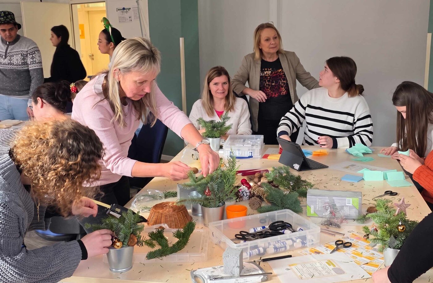Mehrere Personen sitzen und stehen an einem großen Tisch und gestalten gemeinsam weihnachtliche Gestecke aus Tannenzweigen, Zapfen und Christbaumkugeln. Auf dem Tisch liegen Bastelmaterialien, Scheren und Dekorationen in einem hellen Raum.