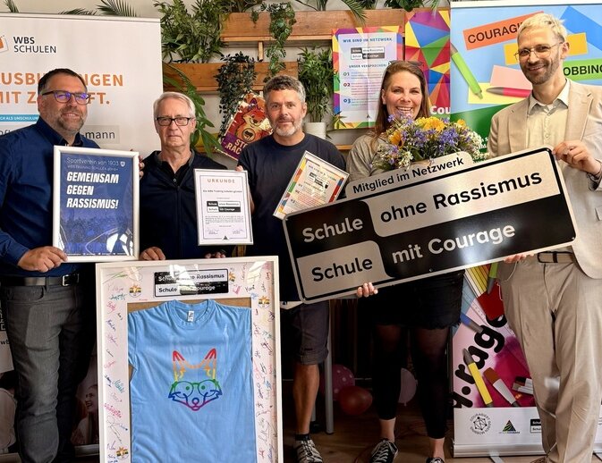 Gruppenfoto bei der Verleihung von „Schule ohne Rassismus – Schule mit Courage“ mit Urkunden, Blumenstrauß und Projektschild.