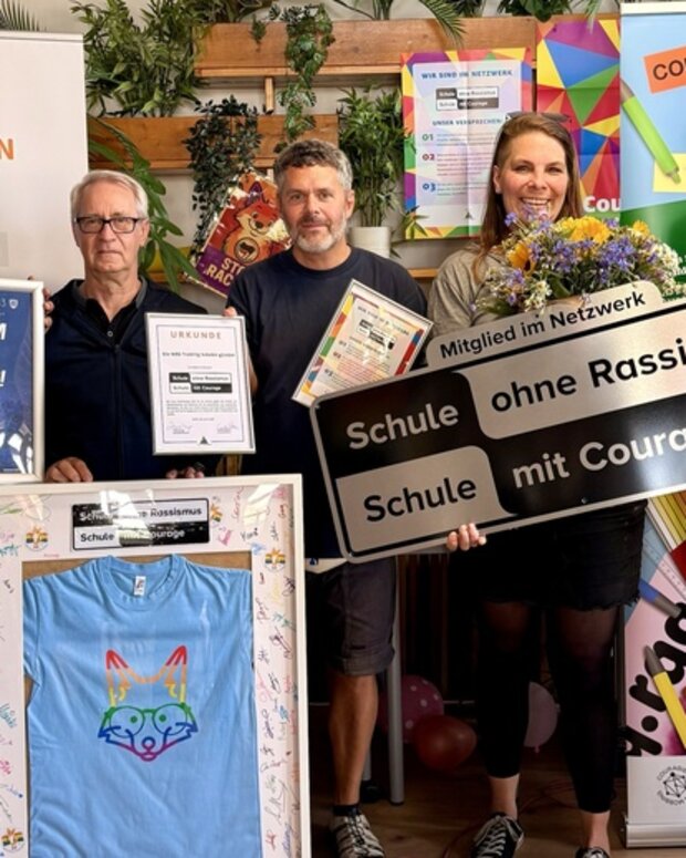 Gruppenfoto bei der Verleihung von „Schule ohne Rassismus – Schule mit Courage“ mit Urkunden, Blumenstrauß und Projektschild.