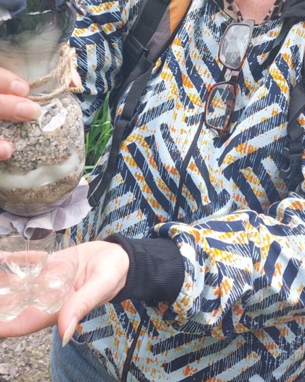 Eine Schülerin demonstriert im Freien einen selbstgebauten Wasserfilter aus einer Kunststoffflasche mit mehreren Filterschichten aus Stoff, Sand und Kies. Gefiltertes Wasser wird in einem transparenten Auffanggefäß gesammelt.
