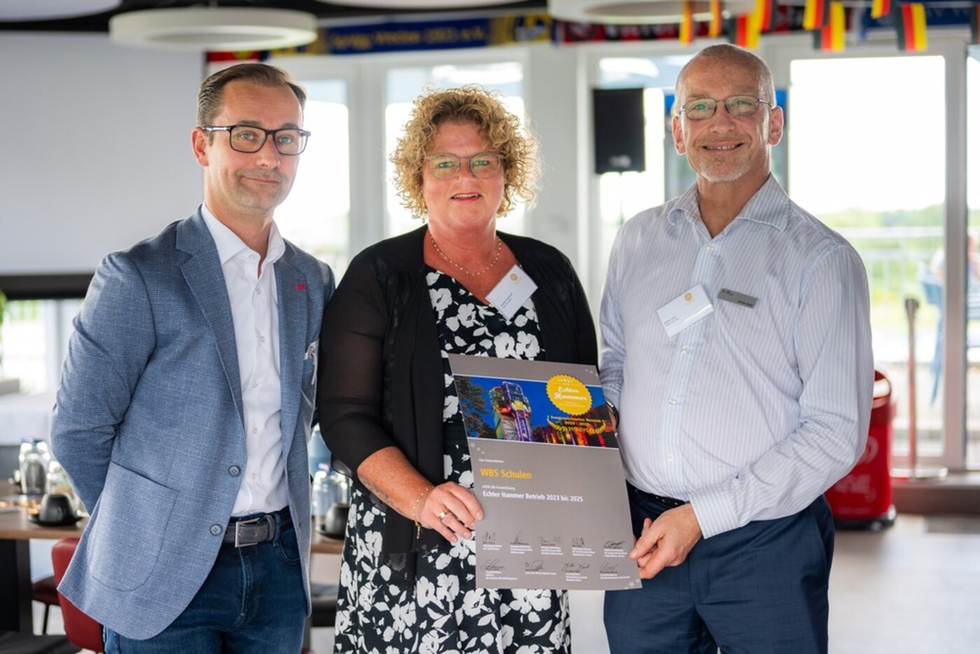 Mitarbeitende der WBS SCHULEN und offizielle Vertretungen der Initiative „Echter Hammer Betrieb“ präsentieren gemeinsam die Auszeichnungsurkunde „Echter Hammer Betrieb“ bei einer Veranstaltung. Die Urkunde wird in die Kamera gehalten.